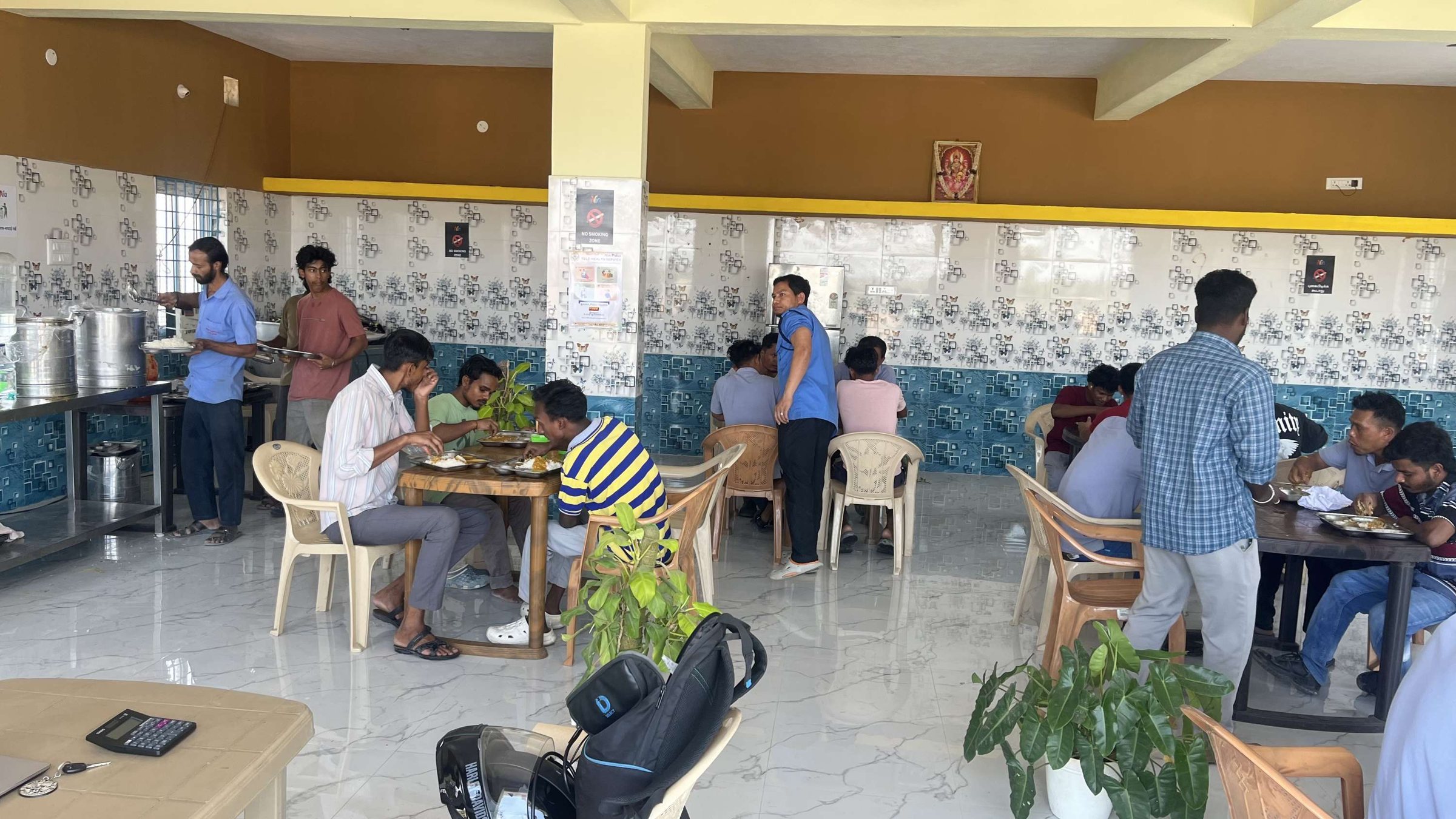Members eating lunch at tables in a bright Nia Studio dining hall while staff serve rice from steel pots